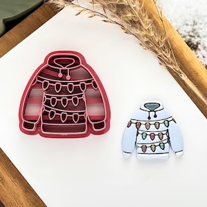 May include: A red plastic cookie cutter and a light blue enamel pin in the shape of a hooded sweater decorated with Christmas lights. The cookie cutter is on the left, and the enamel pin is on the right.