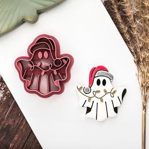 May include: A red plastic ghost-shaped cookie cutter with a Santa hat and string lights. Beside it is a decorated ghost biscuit with a Santa hat and string lights. The biscuit is white with black accents.