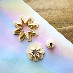 May include: Three cookie cutters, two star-shaped and one flower-shaped, made of beige plastic. The star-shaped cutters have a layered design, with the larger star having a smaller star inside it.