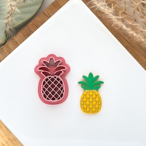 May include: Two pineapple-shaped items. One is a pink cookie cutter with a detailed pineapple design. The other is a yellow and green pineapple-shaped decoration. Both are on a white surface.
