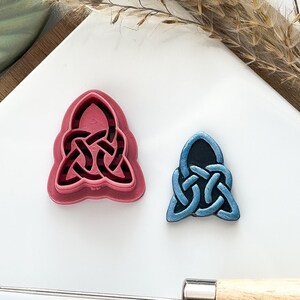 May include: Two Celtic knot-shaped cookie cutters. One is a larger, pink cutter, and the other is a smaller, blue cutter. A metal tool with a wooden handle is in the foreground. The cutters are on a white surface.