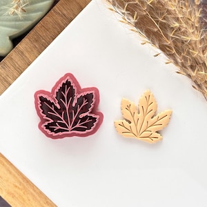 May include: Two leaf-shaped cookie cutters. One is a dark pink with a black interior, and the other is a light tan color. The cutters are on a white surface, with a wooden board and decorative dried plants in the background. The leaf cutters are likely for baking.