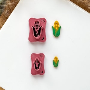 May include: Two pink corn-shaped stamps with the numbers 15 and 10, and two small corn-shaped embellishments. The stamps have a corn graphic and a crescent moon. The corn embellishments are yellow and green.