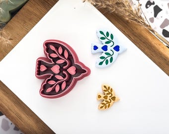 May include: Three bird-shaped objects on a white surface. One is a dark red cookie cutter with leaf and heart details. Another is a white bird with green leaves and blue hearts. The third is a small, tan bird with leaf details.