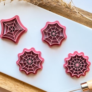 May include: Four pink plastic cookie cutters shaped like spiderwebs. The cutters are on a white surface.