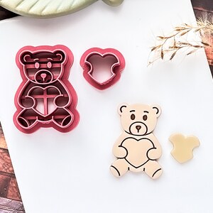 May include: A set of pink teddy bear cookie cutters and a bear-shaped biscuit with a heart. The cutters are in the shape of a teddy bear holding a heart. The biscuit is beige with brown details.