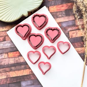 May include: A collection of pink heart-shaped cookie cutters in various designs, including solid hearts and outlines. They are arranged on a white surface, with a decorative green plate and dried ornamental grass in the background.