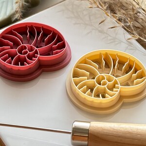 May include: Two seashell-shaped cookie cutters. One is red, the other is yellow. The cutters have a spiral design and are made of plastic. A metal tool with a wooden handle is in the foreground.