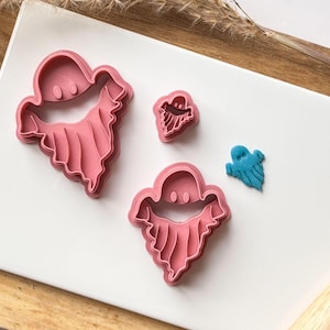 May include: Three pink plastic ghost-shaped cookie cutters on a white surface. The largest cutter is in the foreground, the medium-sized cutter is in the background, and the smallest cutter is in the lower right corner. The smallest cutter is blue.
