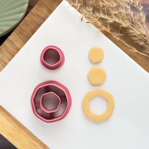 May include: A collection of polymer clay cutters and textured clay shapes in shades of pink and yellow. The cutters are in the shape of a flower and circles. The textured clay shapes are circles and a hexagon. The items are on a white surface.