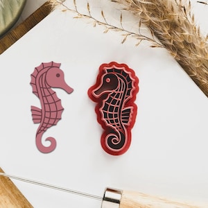May include: Two seahorse-shaped cookie cutters. One is a solid mauve colour, and the other is red with a black interior. The cutters are on a white surface with a wooden cutting board and a metal tool in the background. Decorative dried wheat is also present.