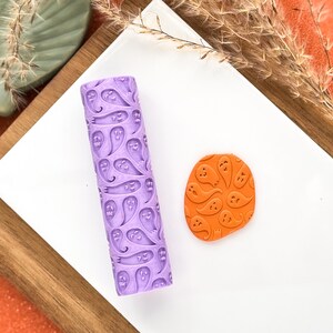 May include: A lilac rolling pin featuring a ghost pattern, alongside an orange clay shape with the same ghost design. The items are on a white surface, with a wooden tray and orange background.