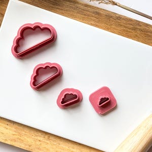 May include: Four pink plastic cookie cutters shaped like clouds. The cutters are arranged in a row on a white surface. The largest cutter is at the top of the image, and the smallest cutter is at the bottom.