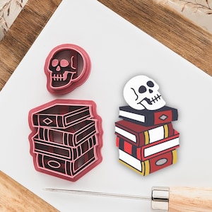 May include: Two pink cookie cutters, one shaped like a skull and the other shaped like a stack of books with a skull on top.