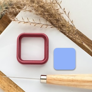 May include: A red plastic square cookie cutter, a light blue square, and a wooden tool with a metal tip on a white surface.