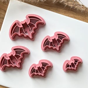 May include: Set of four pink plastic bat-shaped cookie cutters. The cutters are layered, with the largest cutter on top and the smallest cutter on the bottom.