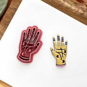 May include: Two hand-shaped decorative items. One is a dark pink outline with a solid pink interior, featuring palmistry symbols. The other is gold with black lines and colourful accents, also with palmistry symbols. Both are on a white surface.