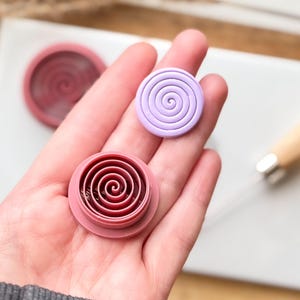 May include: Two round polymer clay molds with a spiral design. One mold is pink and the other is purple.