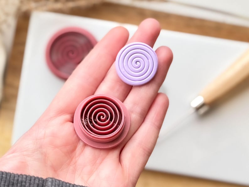 May include: Two round polymer clay molds with a spiral design. One mold is pink and the other is purple.