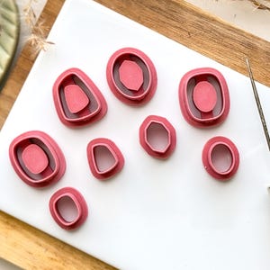 May include: A set of eight pink plastic cookie cutters shaped like different gemstones. The cutters are arranged on a white surface with a wooden background.