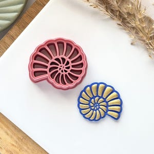 May include: Two seashell-shaped items. One is a pink cookie cutter, and the other is a blue and gold decorative piece. They are on a white surface with a wooden tray and dried plant accents.