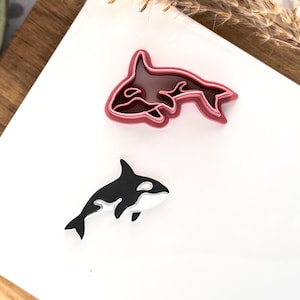 May include: Two orca-shaped items on a white surface. One is a black and white orca figurine. The other is a pink orca-shaped cookie cutter. The items are likely for baking or crafting, with a marine life theme.