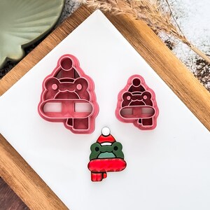 May include: Three frog-shaped cookie cutters. Two are solid pink plastic, one large and one small. The third is a painted frog with a red scarf and hat. The frog is green, red, and white.