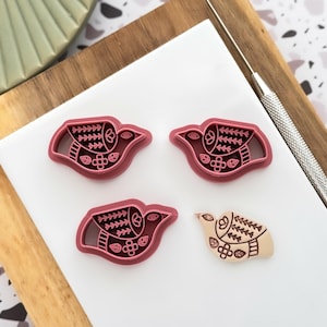 May include: Four bird-shaped cookie cutters and a baked cookie are displayed on a wooden cutting board. The cutters are a deep pink colour and feature intricate designs. A metal tool is also present, suggesting a crafting or baking theme.