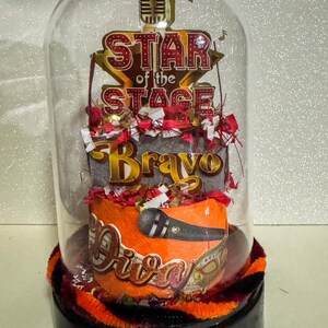 May include: A decorative glass dome featuring a stage-themed design. The dome showcases the words "Star of the Stage", "Bravo", and "Diva" with a microphone graphic. The base is black with orange and black trim.