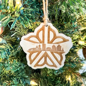 May include: A wooden snowflake ornament with a laser-cut design featuring a cityscape silhouette. The ornament is hanging on a green Christmas tree.