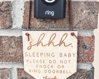 May include: A wooden sign with the text "shhh. Sleeping baby please do not knock or ring doorbell thank you" hangs on a brick wall next to a black Ring doorbell.