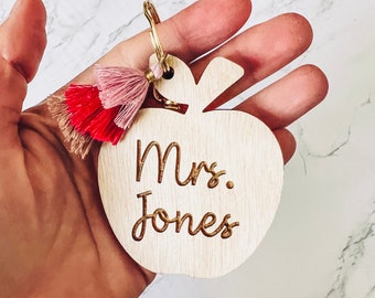 Puede incluir: Llavero de madera con forma de manzana con un borla en tonos rosa y rojo. El llavero está grabado con el texto "Mrs. Jones".
