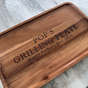 May include: A rectangular wooden serving tray with rounded corners and a natural grain finish. The surface is laser-engraved with the text POP'S GRILLING PLATE and the inscription Happy Father's Day 2026 in a dark, clear, sans-serif font.