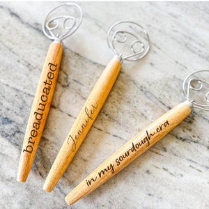 Puede incluir: Tres batidores de masa de madera con bucles de alambre de metal. Los batidores est&aacute;n grabados con el texto "breadeducated", "Jennifer" y "in my sourdough era".