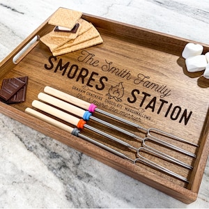 May include: Wooden tray with the text "The Smith Family Smores Station" and a campfire graphic. The tray includes graham crackers, chocolate bars, marshmallows, and four metal skewers with wooden handles.