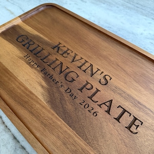 May include: A rectangular wooden serving tray with a raised edge and natural grain patterns. The surface is laser-engraved with the text KEVIN'S GRILLING PLATE and Happy Father's Day 2026 in a dark, bold, sans-serif font centered on the wood.