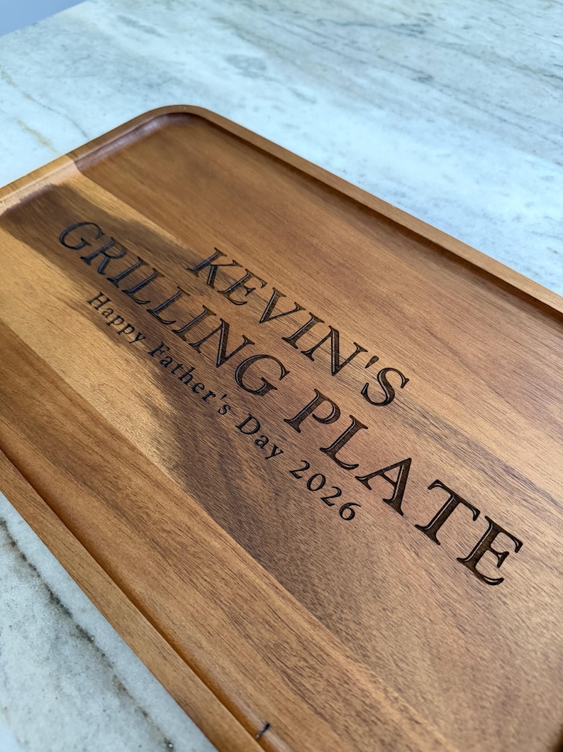 May include: A rectangular wooden serving tray with a raised edge and natural grain patterns. The surface is laser-engraved with the text KEVIN'S GRILLING PLATE and Happy Father's Day 2026 in a dark, bold, sans-serif font centered on the wood.