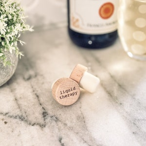 May include: A wooden bottle stopper with the text "liquid therapy" engraved on it. The stopper is on a white marble surface.