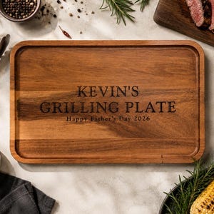 May include: A rectangular wooden serving tray with rounded corners and a grooved border. The center is engraved with the text KEVIN'S GRILLING PLATE and Happy Father's Day 2026 in a dark, serif font, showcasing the natural brown wood grain.