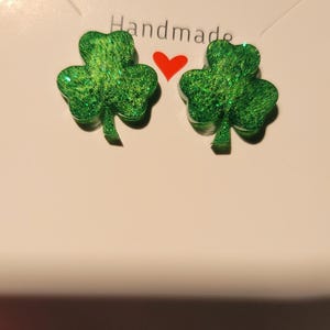 May include: Handmade green glitter shamrock earrings. The earrings are in the shape of a three-leaf clover, with a red heart between them. The word "Handmade" is above the earrings.