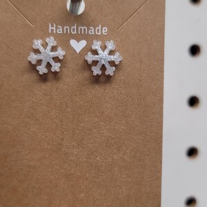 May include: A pair of handmade snowflake earrings with a glittery finish. The earrings are displayed on a brown card with the word "Handmade" printed on it. The earrings are a winter-themed accessory.