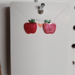 May include: Red apple-shaped earrings with green leaves, displayed on a white card. The word "Handmade" is visible on the card. The earrings are attached to a pegboard.