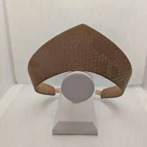May include: A tan headband with a snakeskin pattern. The headband has a wide, curved design with a pointed top. It is displayed on a white stand against a white background.