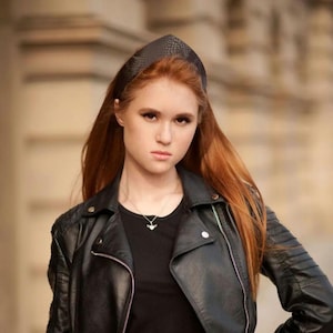 Può includere: Una donna con lunghi capelli rossi indossa una giacca di pelle nera e una fascia nera strutturata. La giacca ha una chiusura a zip. Indossa anche una collana d'argento. Lo sfondo è sfocato.