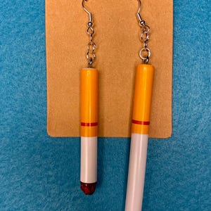Op de afbeelding: Een paar oorbellen in de vorm van sigaretten. Elke oorbel heeft een witte behuizing, een oranje bovenkant en een rode band. De oorbellen hangen aan zilverkleurige oorhaakjes.