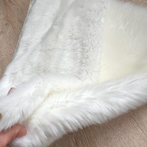 May include: A white faux fur rug with a soft, fluffy texture. The rug is folded in half, showing the underside of the fur.