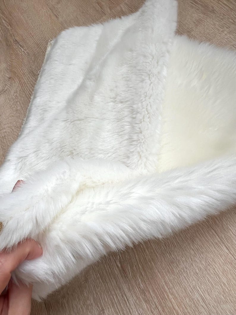 May include: A white faux fur rug with a soft, fluffy texture. The rug is folded in half, showing the underside of the fur.