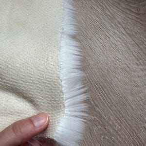May include: Close-up of a beige fabric with a white faux fur trim. The fabric is being held in a hand.