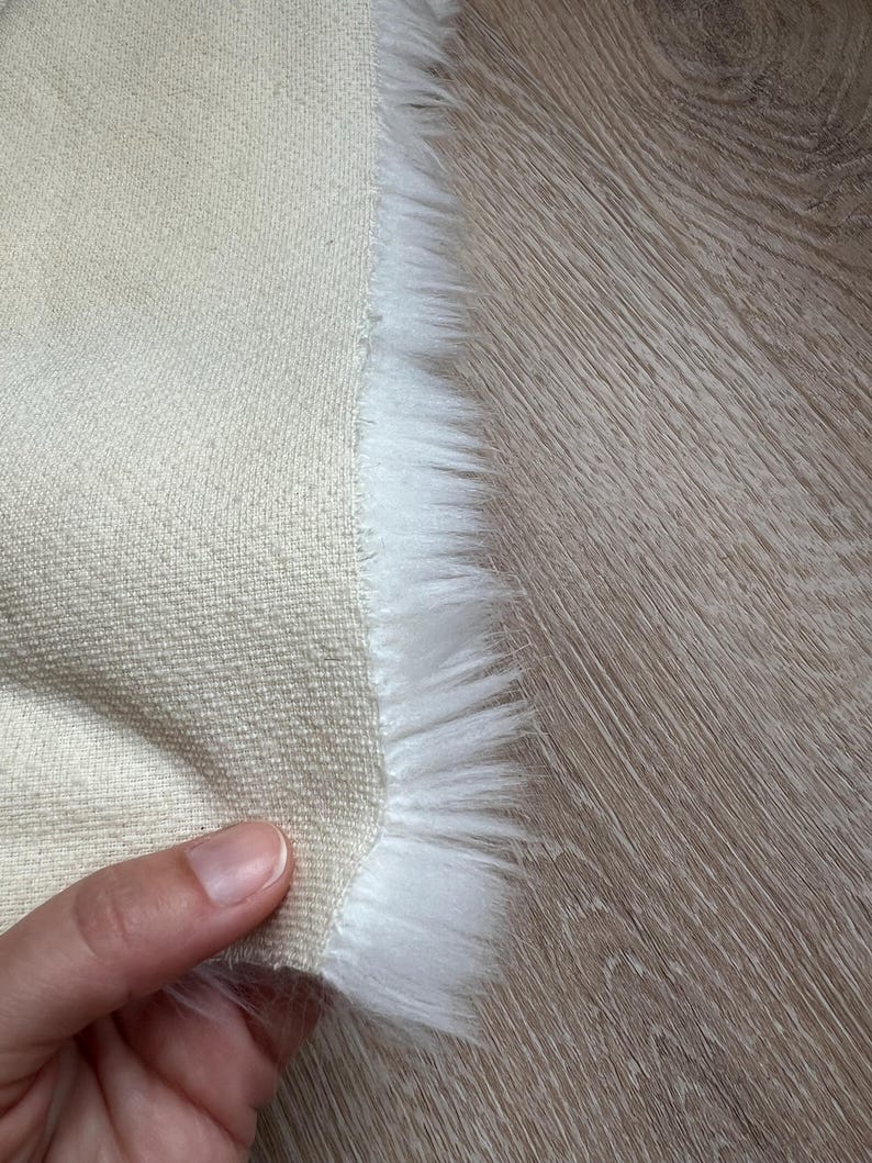 May include: Close-up of a beige fabric with a white faux fur trim. The fabric is being held in a hand.