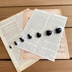 Könnte beinhalten: Eine Sammlung schwarzer Bärenaugen und weißer Unterlegscheiben sowie schwarzer Sicherheitsaugenschrauben. Die Bastelmaterialien sind auf Vintage-Papier mit gedrucktem Text und Noten angeordnet, vor einem Holzgrund.